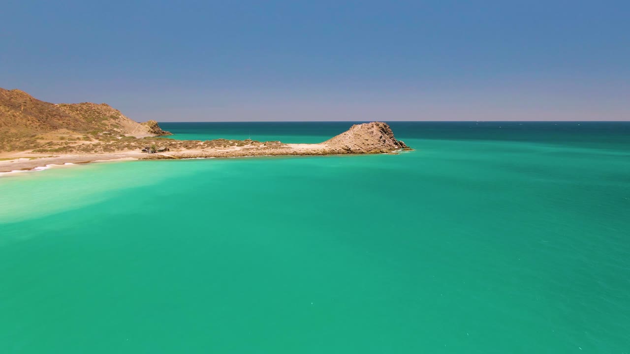 Aerial frame at 0:02 over Cabo Pulmo with green-blue water across the foreground, the pale beach edge at left, dry hills behind it, the rocky point slightly right of center, and no visible people.