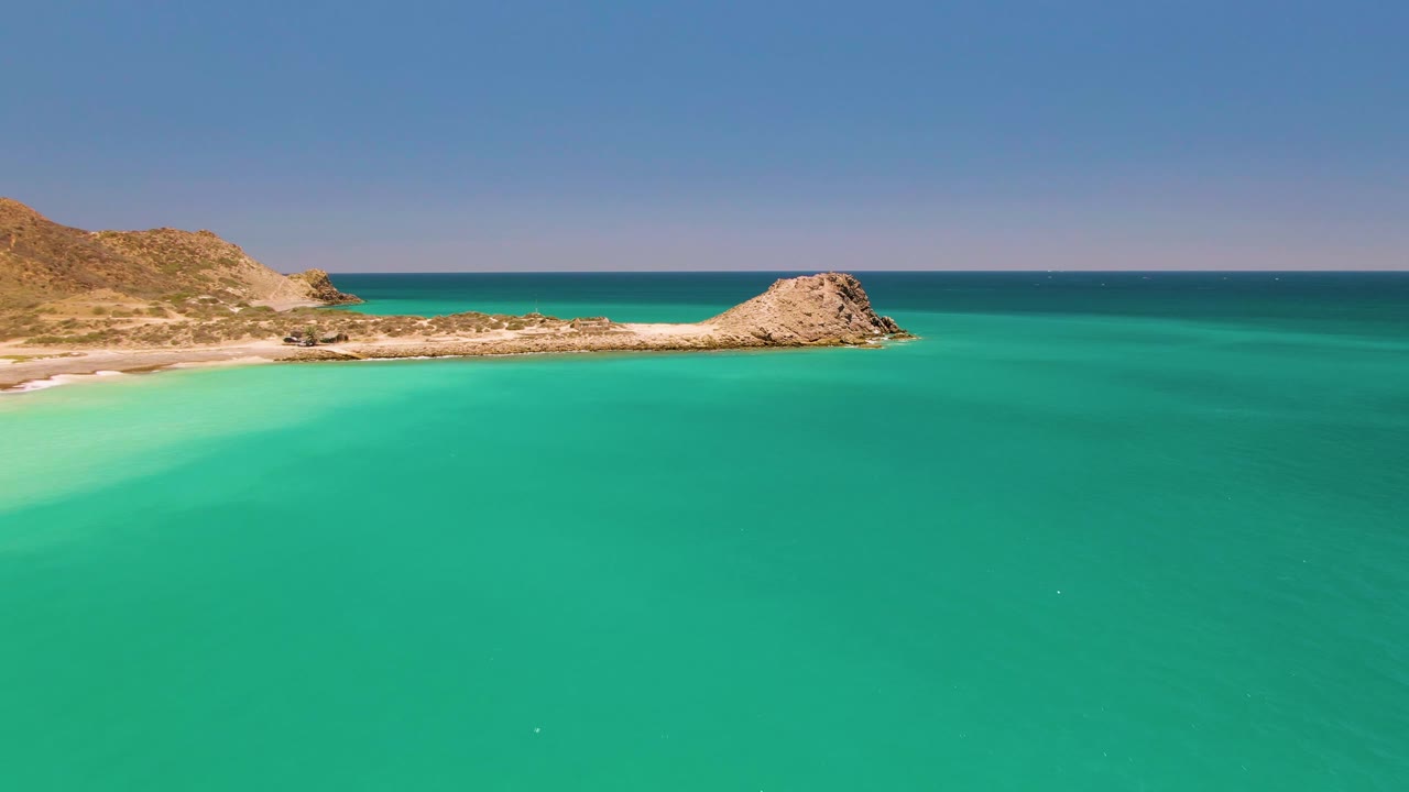 Mid-clip aerial frame over Cabo Pulmo showing shallow turquoise water close to the beach, the rocky hillside entering from the left, the low outcrop along the shoreline, and no visible people.
