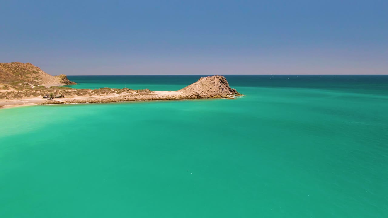 Wide aerial frame of Cabo Pulmo coastline with turquoise water in the foreground, a sandy strip and rocky peninsula across the middle, dry hills to the left, and no visible people.