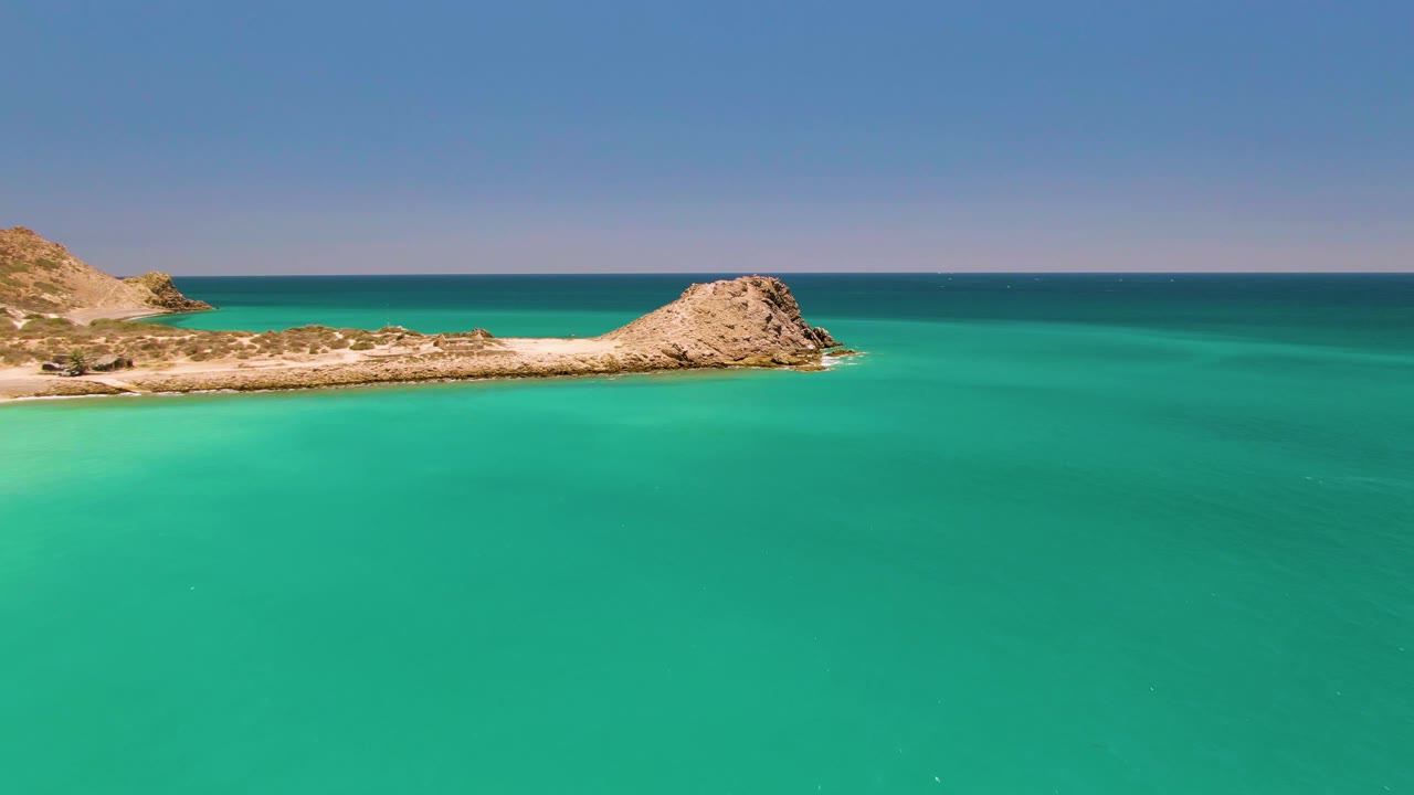 Aerial Cabo Pulmo frame with the rocky peninsula stretching across the upper third, shallow turquoise water below, darker ocean beyond, and no visible people.