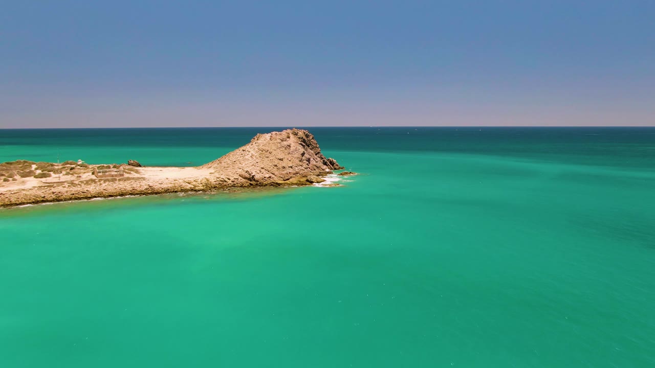 Aerial frame of the Cabo Pulmo rocky islet set farther across the top of the image, with calm turquoise water filling most of the view and no visible people.