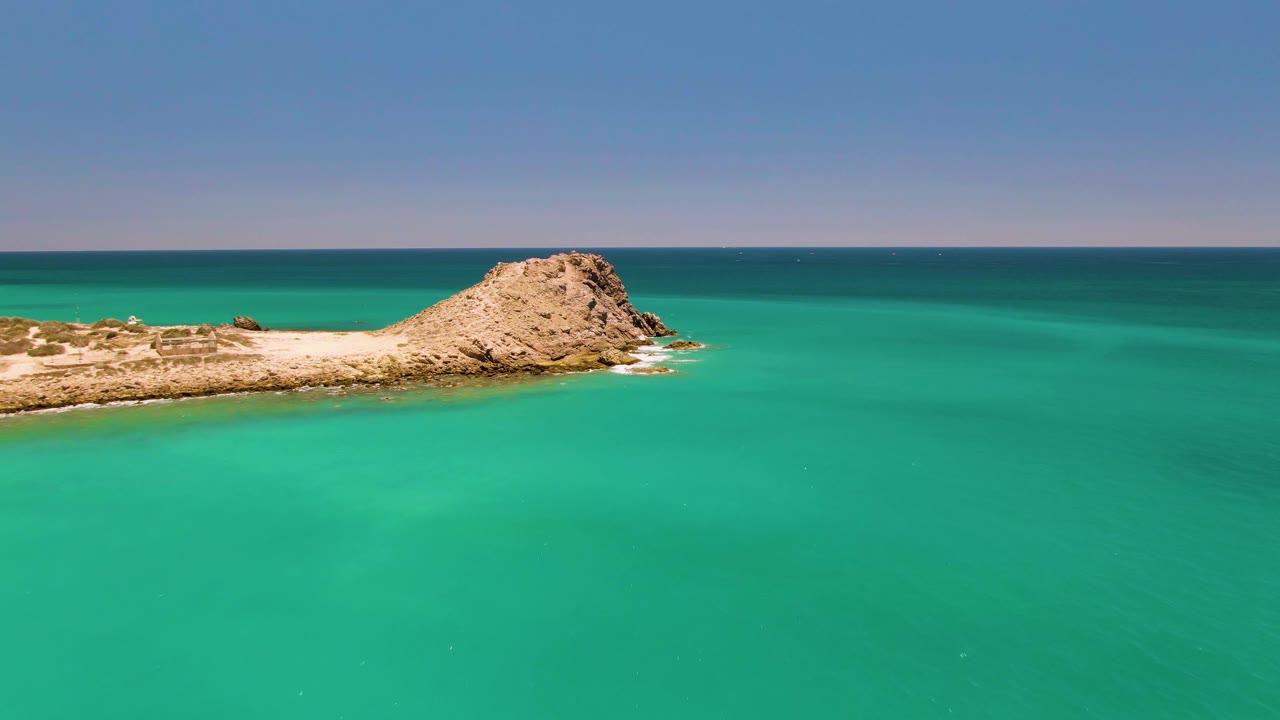 The rocky islet remains near the top of the aerial frame in Cabo Pulmo, with smooth turquoise sea below, a dark blue horizon line, and no visible people.