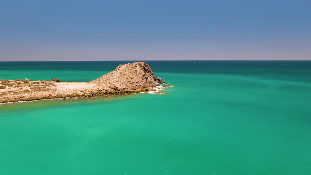 Aerial view of the Cabo Pulmo islet shifted lower and left in frame, with shallow turquoise water, a flat horizon, clear sky, and no visible people.