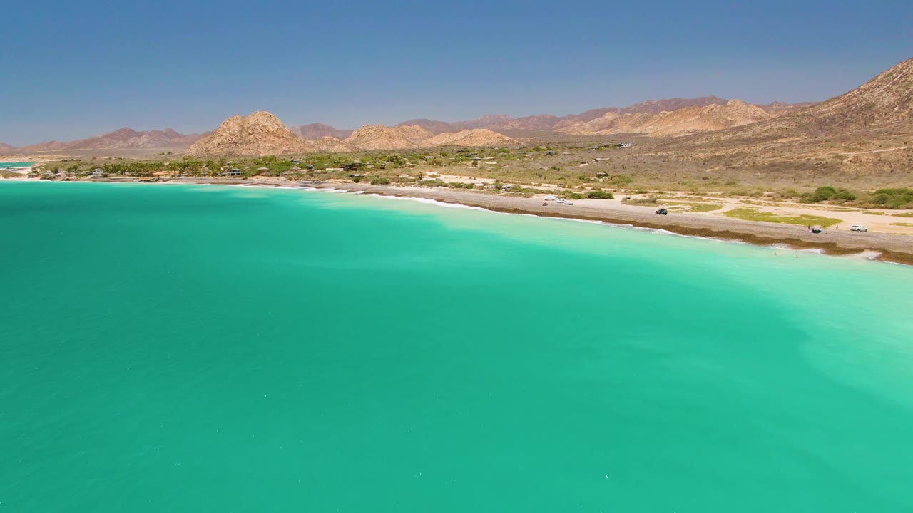 Aerial view of turquoise water beside a sandy coastal road and mountains.