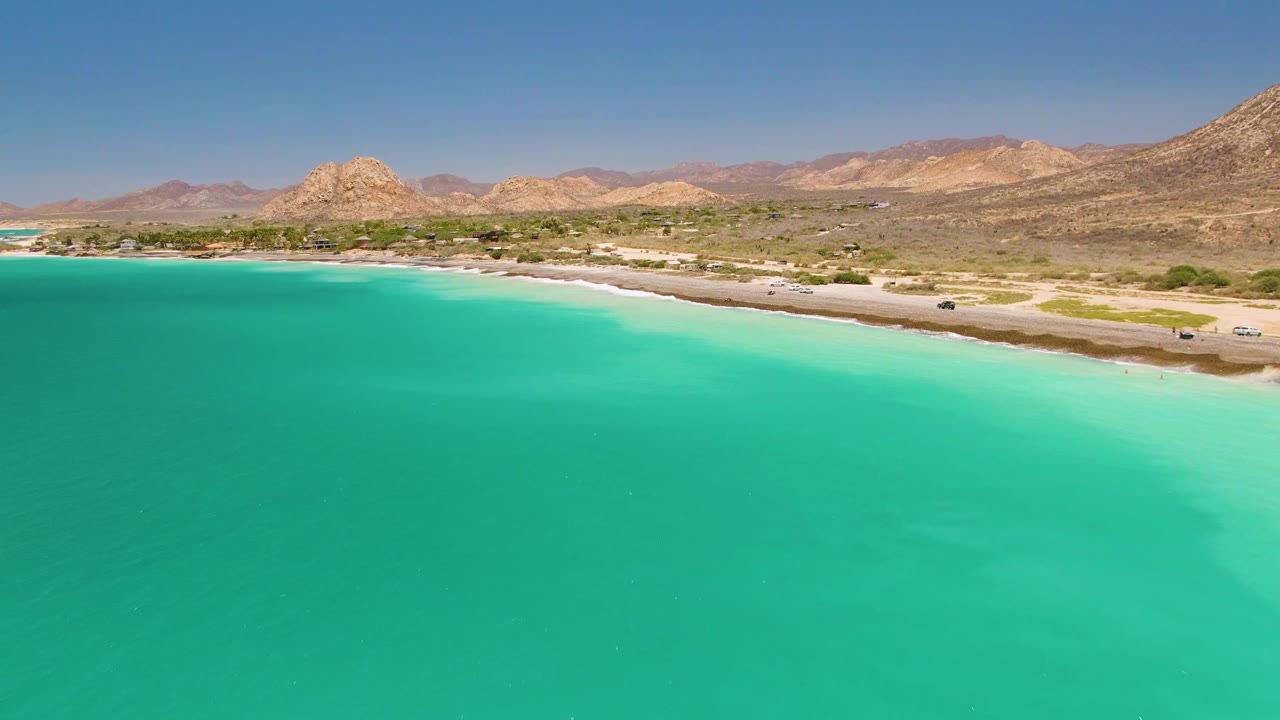 Aerial view in Cabo Pulmo showing broad turquoise water, a pale beach edge, small vehicles along the coastal road, arid hills, distant mountains, and no visible people.