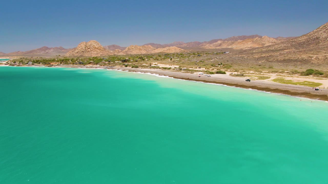 Aerial frame from Cabo Pulmo with the water extending across the foreground, the beach and road crossing the middle, small vehicles near the shore, mountains on the horizon, and no visible people.