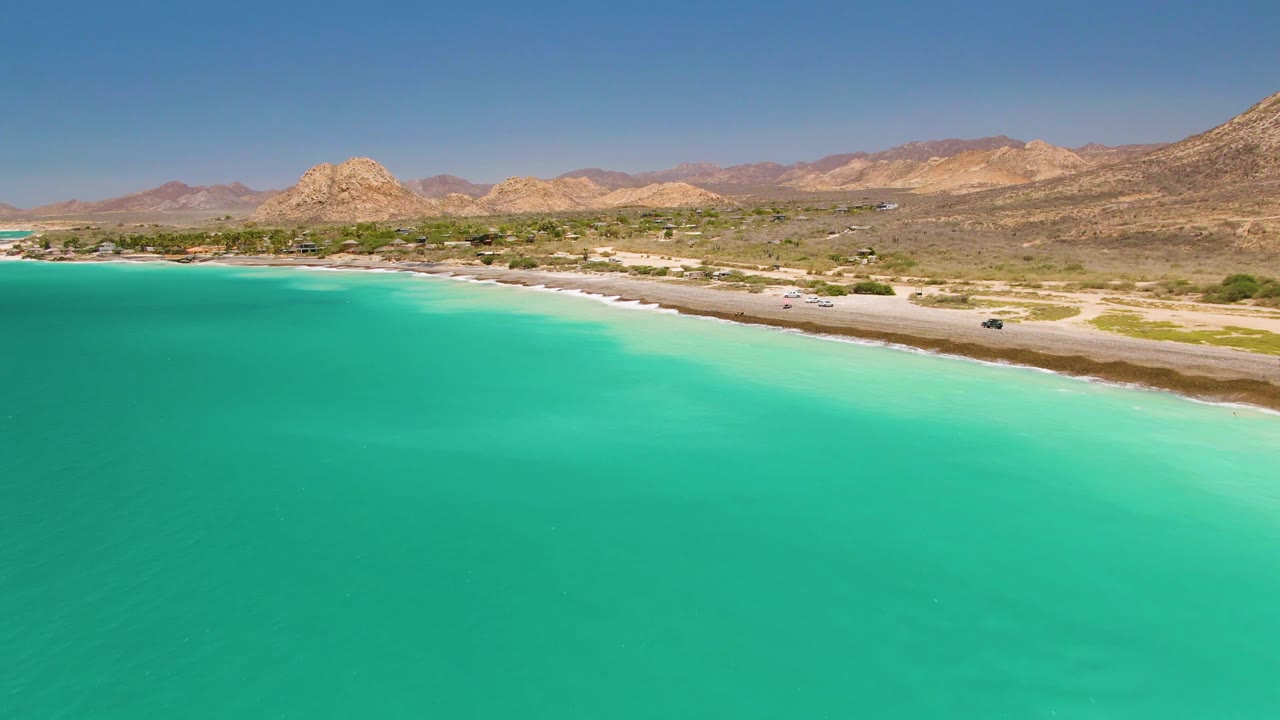 Drone-style aerial view over Cabo Pulmo with turquoise water below, a light sandy coastal road with vehicles, rocky desert mountains beyond, clear sky, and no visible people.