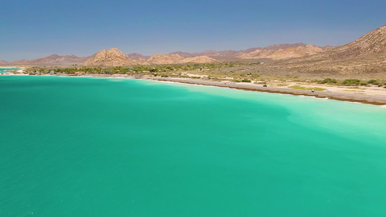 Opening frame of Cabo Pulmo with turquoise water in the foreground, a sandy beach and coastal road in the midground, arid mountains beyond, and no visible people.
