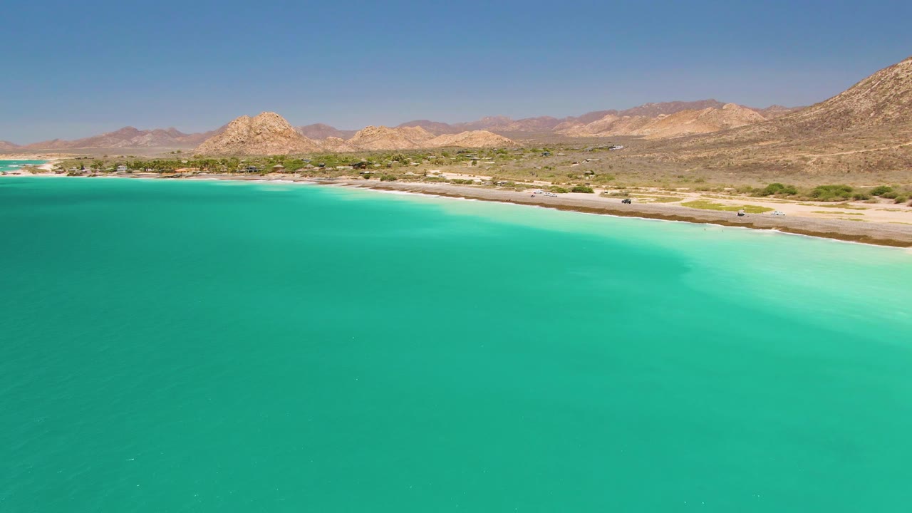 Cabo Pulmo aerial frame with the blue-green ocean taking most of the foreground, the road tracking along the beach, small vehicles on the shore road, and no visible people.