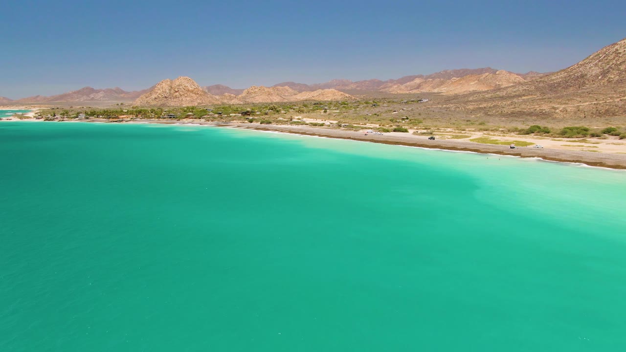 Wide Cabo Pulmo frame showing uninterrupted turquoise water below, a narrow sandy shore and road across the middle, layered dry mountains behind, and no visible people.