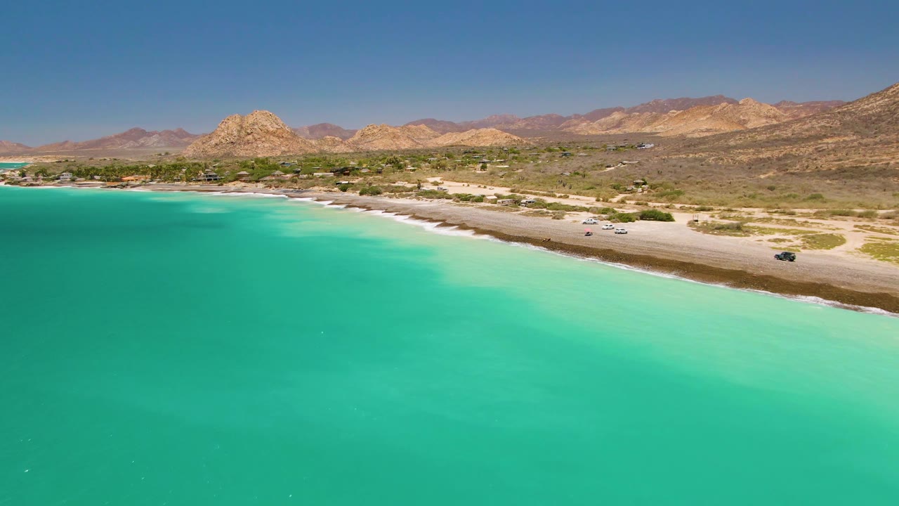 Opening aerial frame over Cabo Pulmo with turquoise water in the foreground, a pale beach road along the shore, arid mountains beyond, and no visible people.