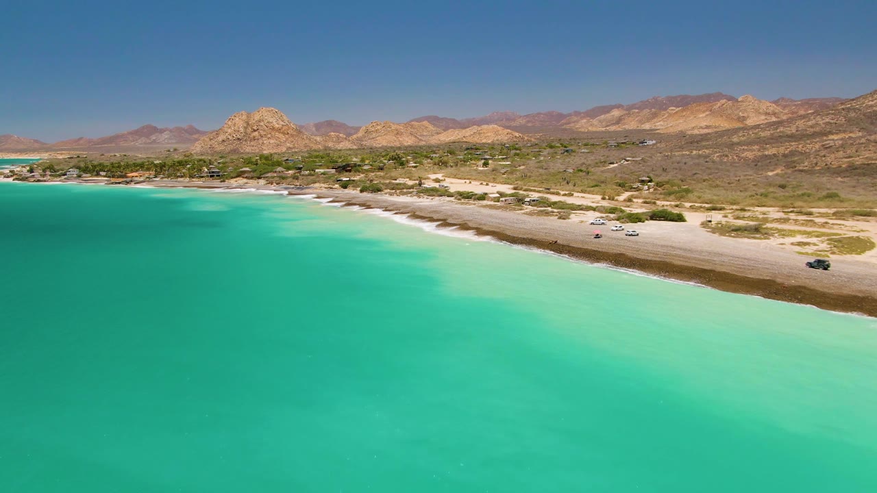 Turquoise shoreline with cars on a sandy road below arid hills.
