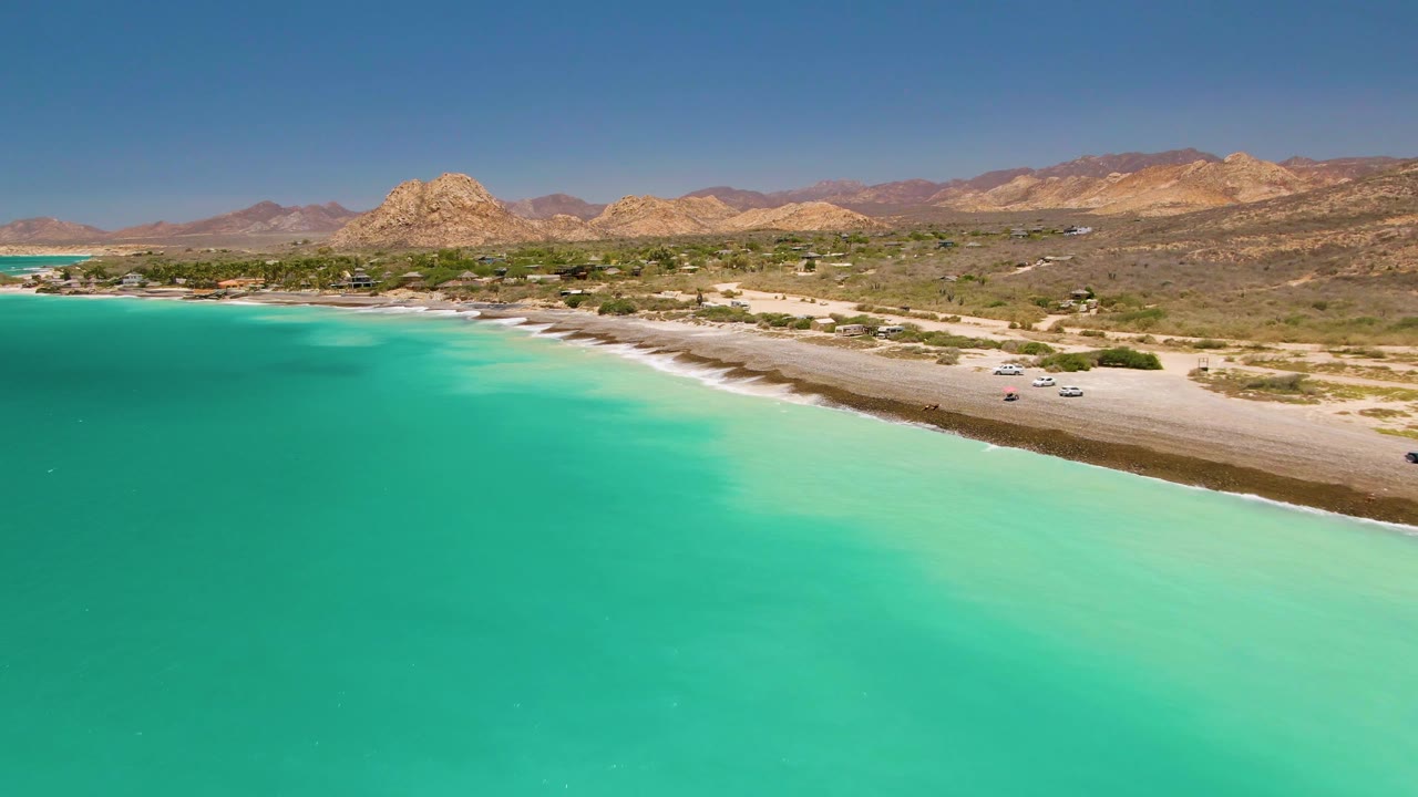 Aerial frame at 0:02 from Cabo Pulmo with broad turquoise water in front, a narrow surf line at the beach road, scattered vehicles, rocky mountains, and no visible people.