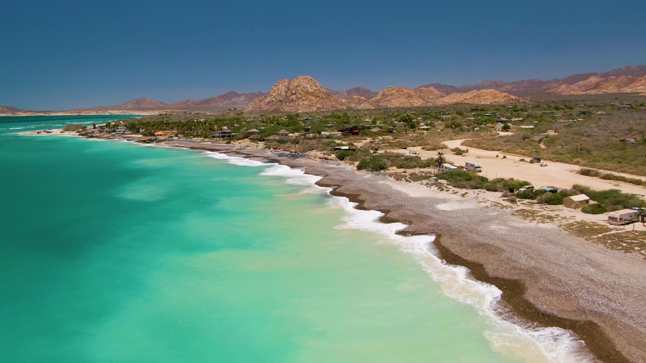 Opening aerial frame over Cabo Pulmo, Baja California Sur, with turquoise ocean in the foreground, a pale shoreline curving right, desert vegetation, distant mountains, and no visible people.