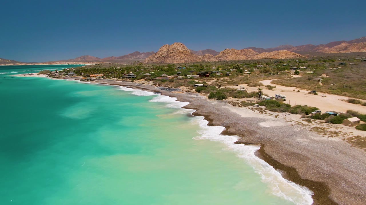 Aerial view one second into the Cabo Pulmo clip, with shallow green-blue water, white wave lines, a longer visible beach edge, scattered small structures, desert terrain, and no visible people.