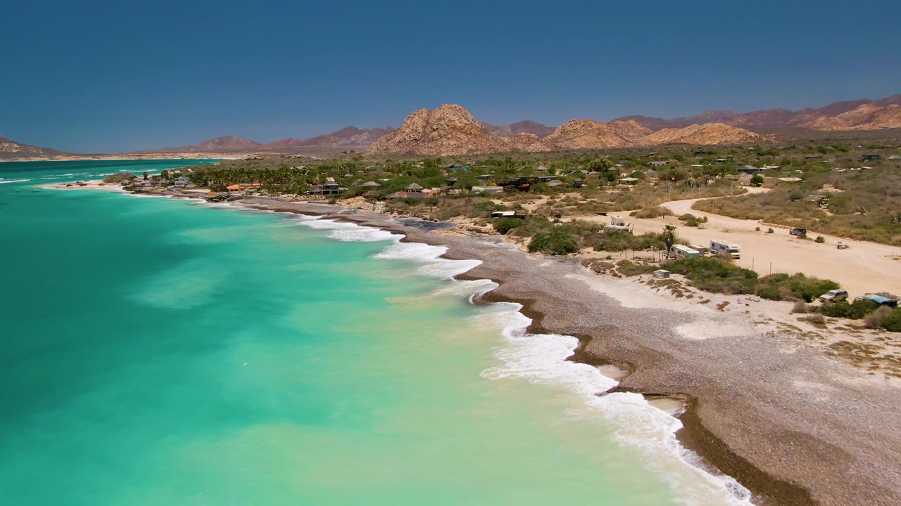 Mid-clip aerial frame from Cabo Pulmo showing turquoise shallows across the left side, foamy surf along the beach, sparse coastal development, dry vegetation, mountains, and no visible people.