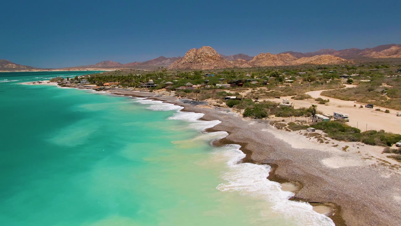 Aerial coastline looking along turquoise sea, repeated surf lines, sandy beach, and desert hills.