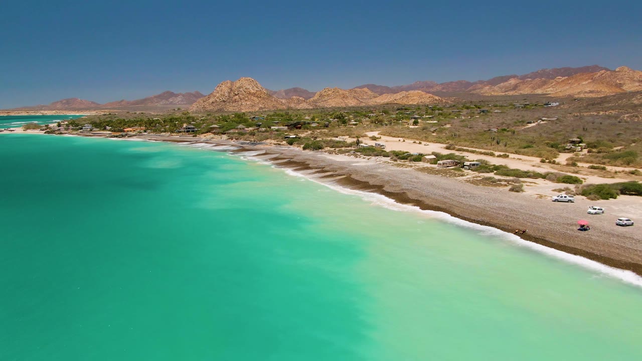 Aerial frame over Cabo Pulmo showing turquoise water in the foreground, a pale sandy and rocky shore with several cars near the beach, dry hills behind it, and no visible people.