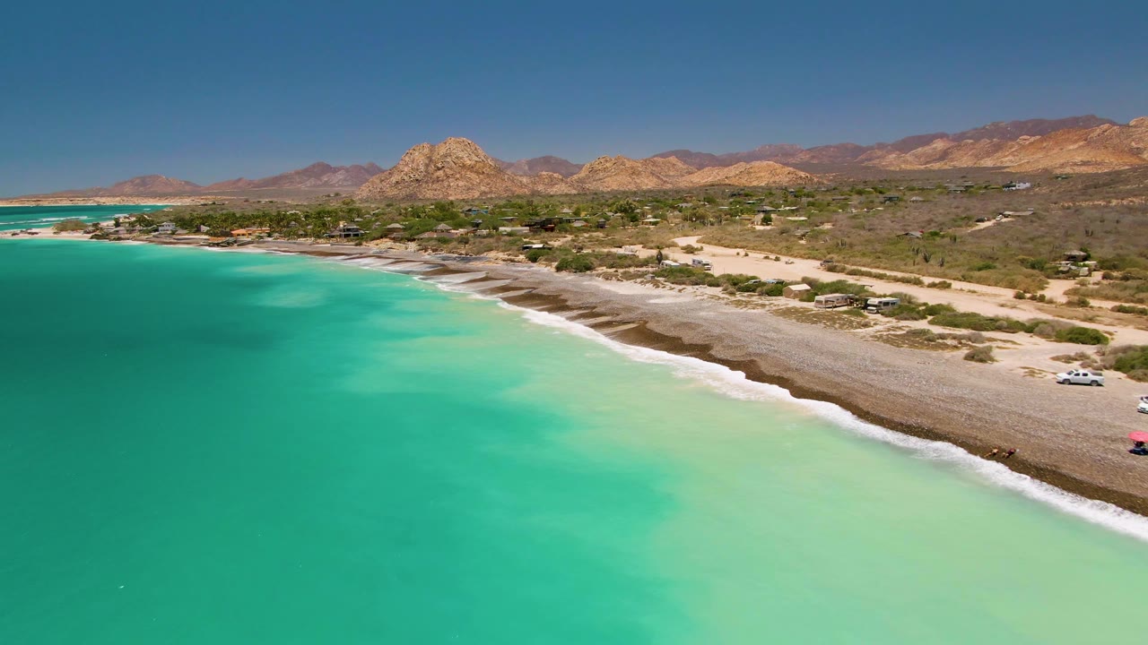 Drone view along a turquoise coastline with cars near the beach and mountains behind.