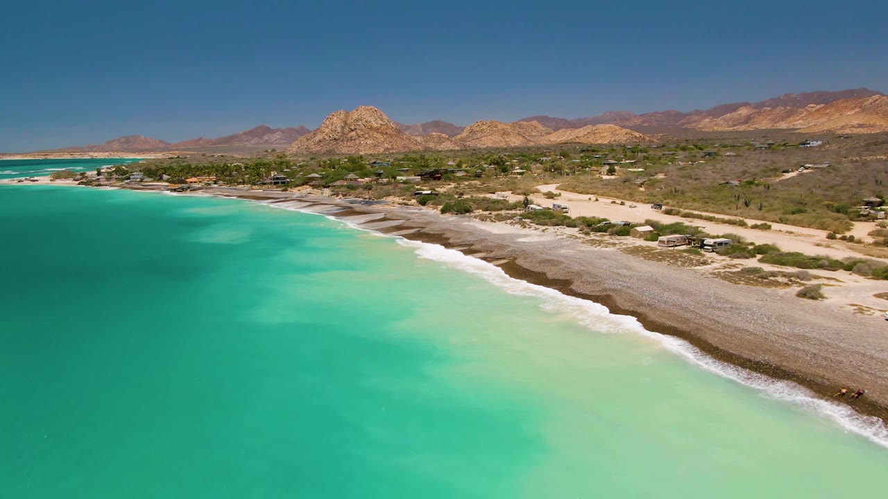 Cabo Pulmo aerial frame with a broad band of pale turquoise shallows, white surf tracing the shoreline, sparse beach structures or shade areas inland, desert hills, and no visible people.
