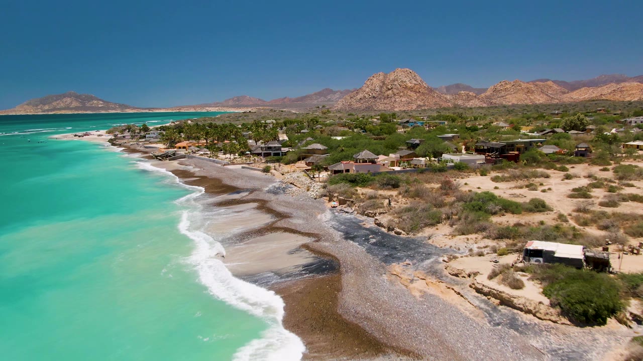 Aerial frame over Cabo Pulmo with turquoise water at left, foamy surf on the shore, beachfront homes and palms in the midground, rocky mountains behind, and no clearly visible people.