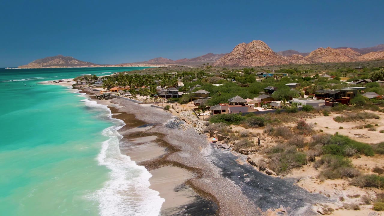 Aerial frame of Cabo Pulmo showing a broad band of turquoise sea, a white wave edge, sandy and rocky beach, low coastal buildings, palms, dry brush, and mountains under a clear sky.