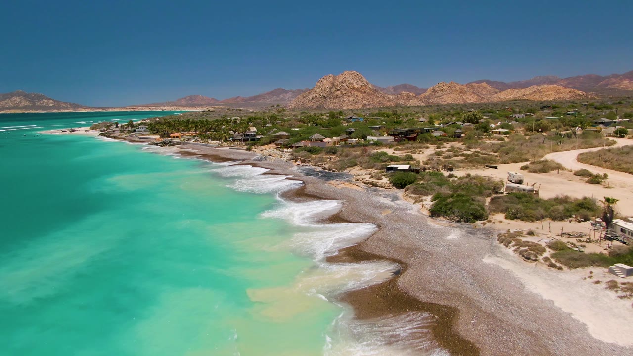 At 0:00, an aerial view of Cabo Pulmo shows turquoise water on the left, white surf reaching a sand and pebble shoreline, dry coastal vegetation, scattered small structures, rocky hills, and no visible people.