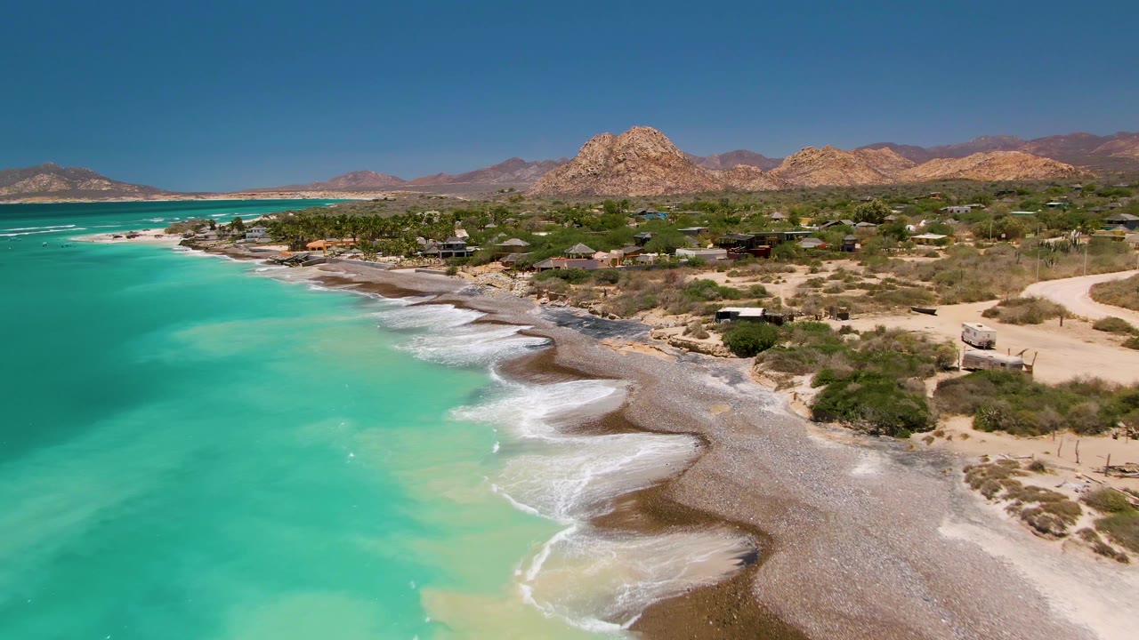 At 0:01, the Cabo Pulmo shoreline curves through the frame as white foam spreads over the beach, turquoise water fills the left side, small structures sit among vegetation, and no visible people appear.