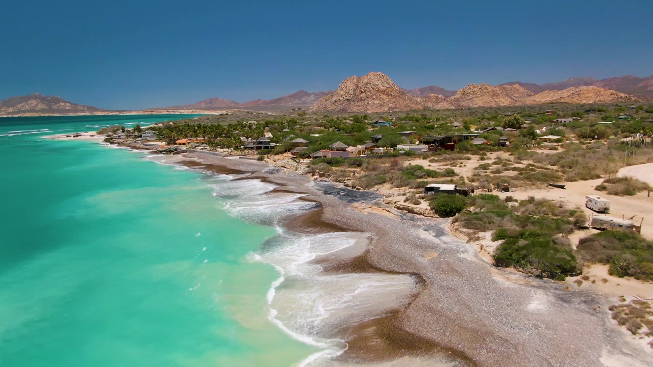 At 0:02, foamy waves mark the beach edge at Cabo Pulmo while the drone view includes turquoise shallows, dry shrubs, low buildings, pale dirt tracks, rocky hills, and no visible people.