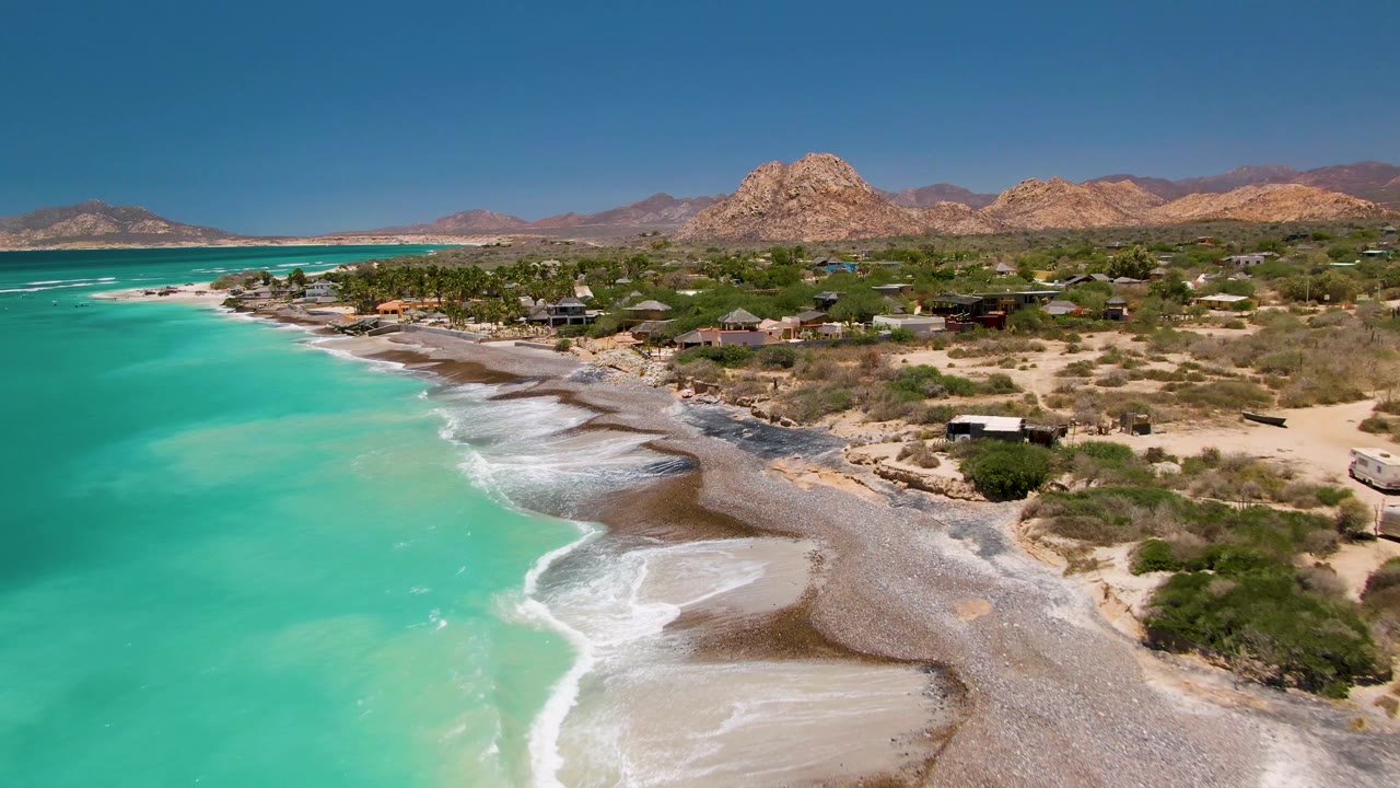 Wide aerial of turquoise sea beside a surf-lined beach and coastal settlement.