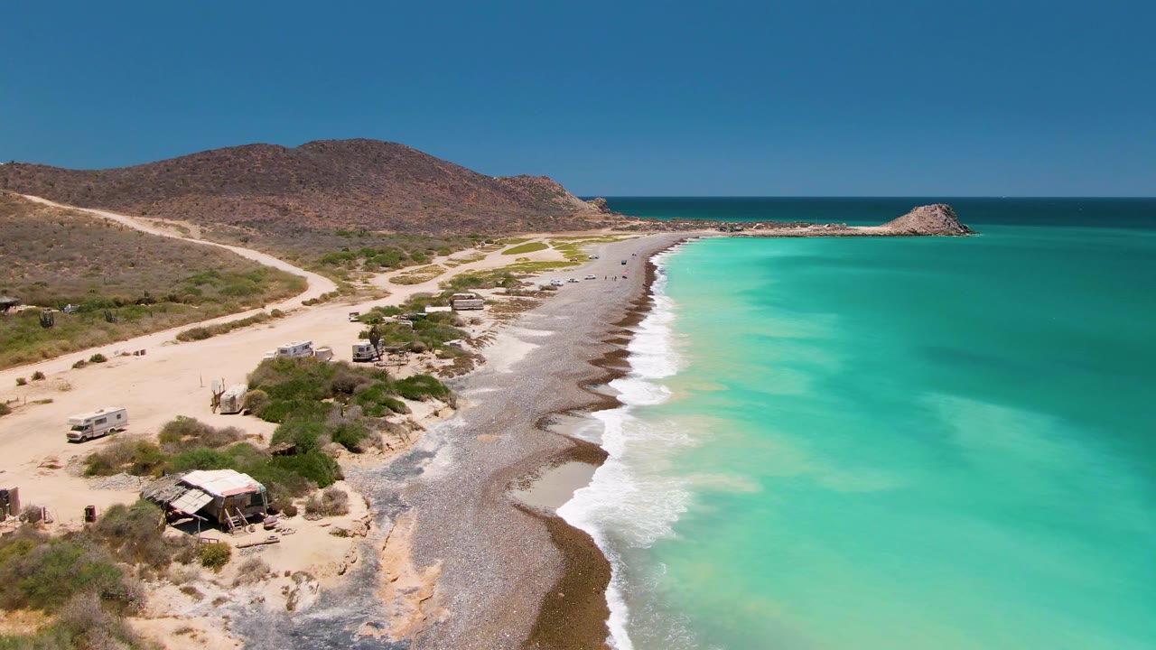Aerial frame over Cabo Pulmo showing a pale dirt road beside a sandy and rocky beach, turquoise water, low scrub, distant brown hills, and no visible people.