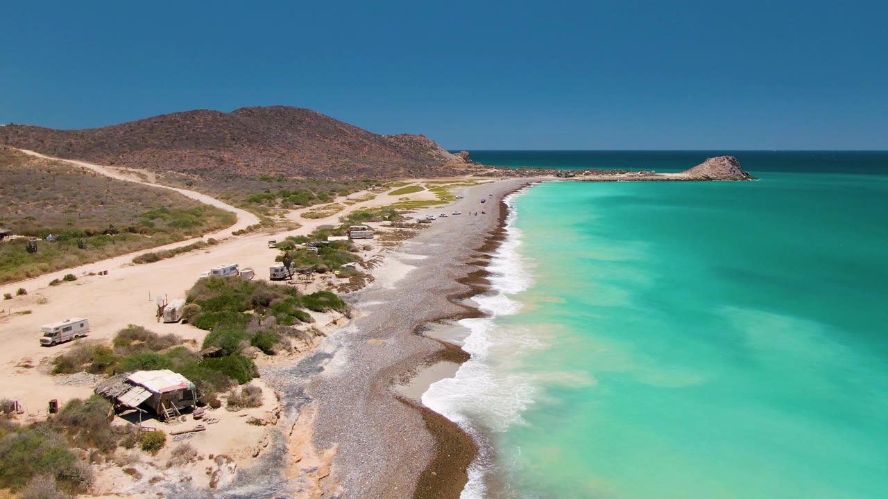 Coastal dirt road with vehicles beside turquoise waves and dry hills.