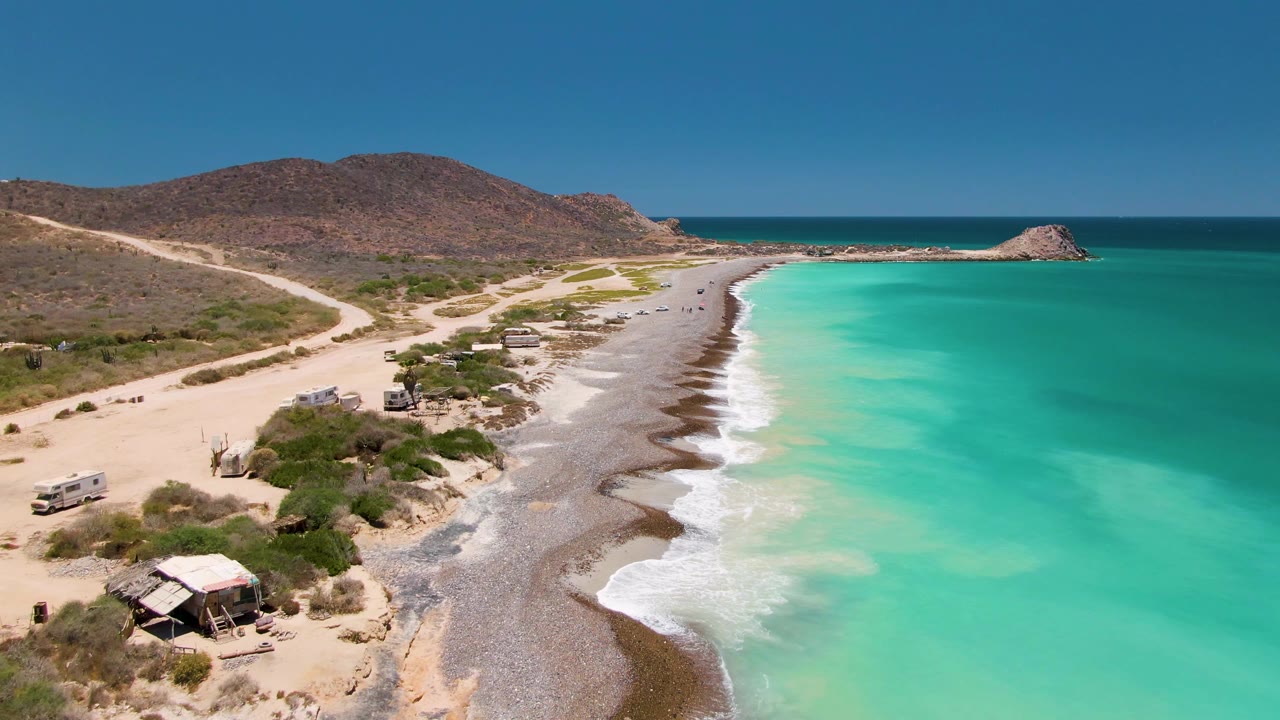 Aerial frame over Cabo Pulmo with the shoreline centered, parked or moving vehicles near the dirt road, turquoise sea spreading across the right side, and no visible people.