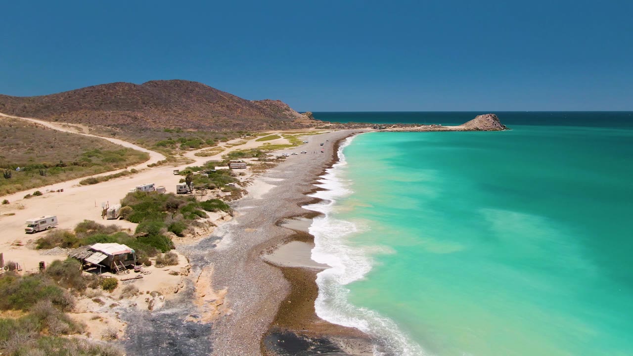 Aerial frame over Cabo Pulmo with turquoise water on the right, white surf along a curved beach, a coastal road with vehicles on the left, dry hills beyond, and no visible people.