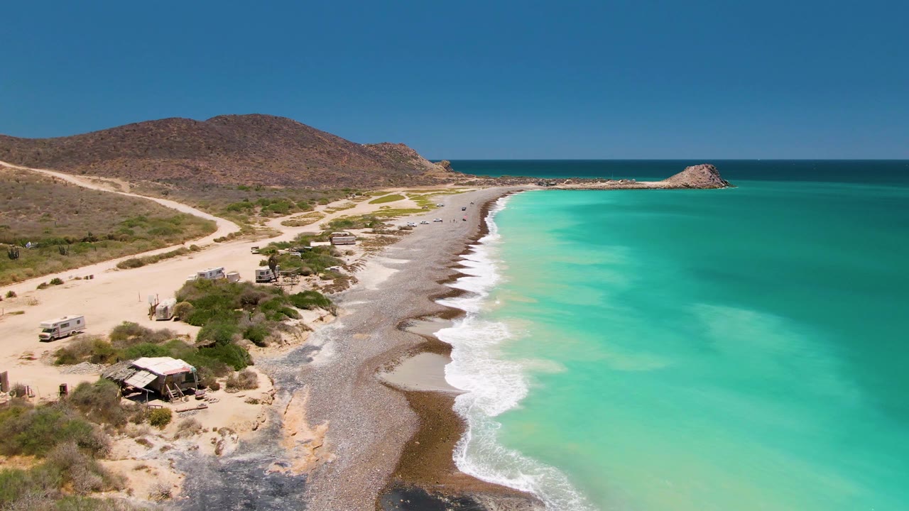 Cabo Pulmo shoreline seen from above with pale surf reaching the beach, vehicles parked near the dirt road, scattered scrub, low desert hills, and no visible people.