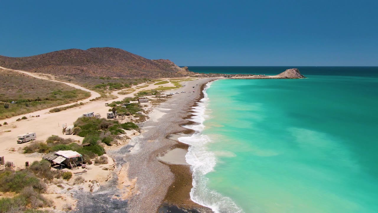 Elevated Cabo Pulmo view with the ocean filling the right side, foam tracing the shoreline, a light-colored road and vehicles to the left, brown hills in back, and no visible people.