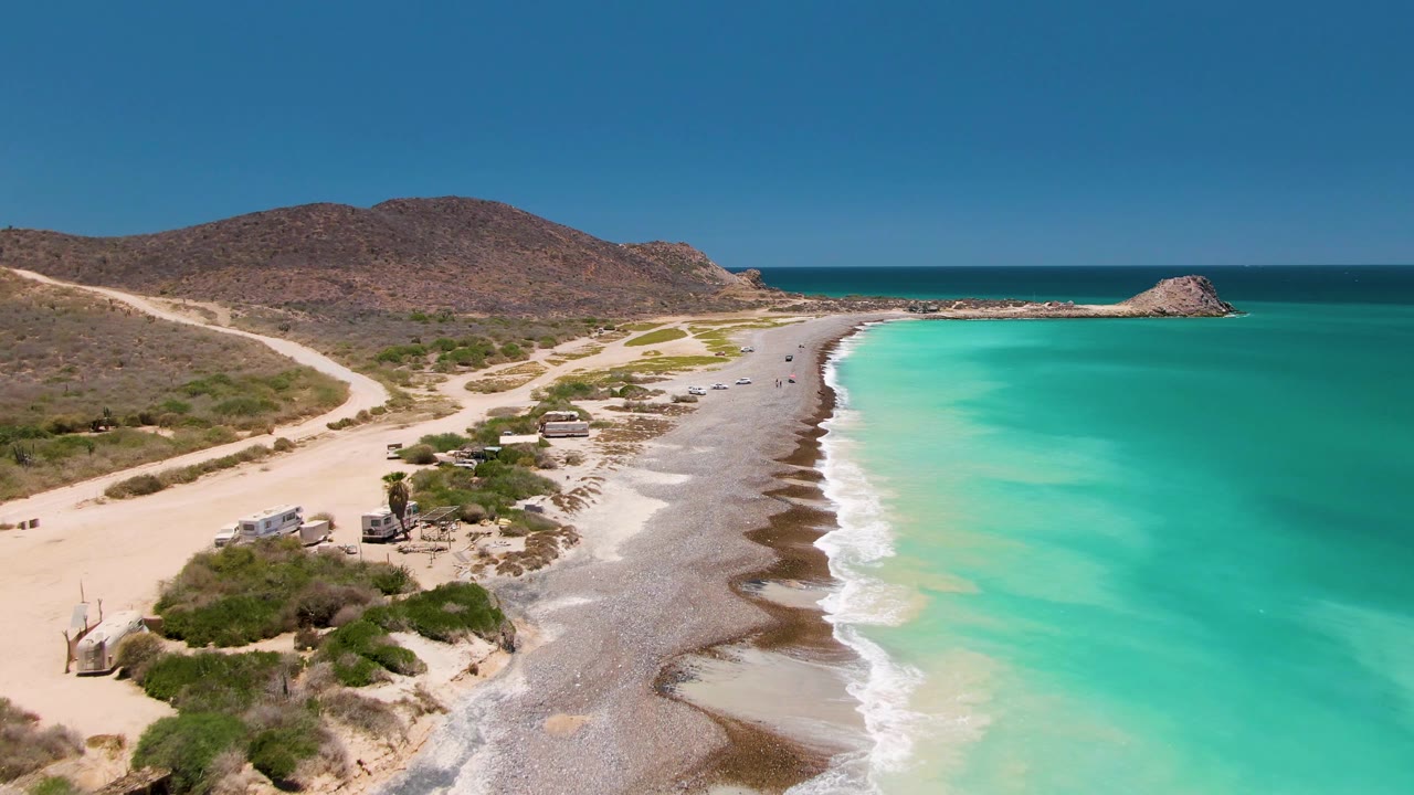 Aerial view in Cabo Pulmo, Baja California Sur, showing a sandy coastal road, parked or moving vehicles, pale beach, turquoise ocean, dry hills, clear sky, and no visible people.