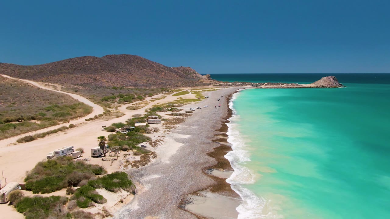 Elevated drone view in Cabo Pulmo with the sandy road and vehicles lower left, a narrow beach through the center, turquoise water to the right, dry hills beyond, and no visible people.
