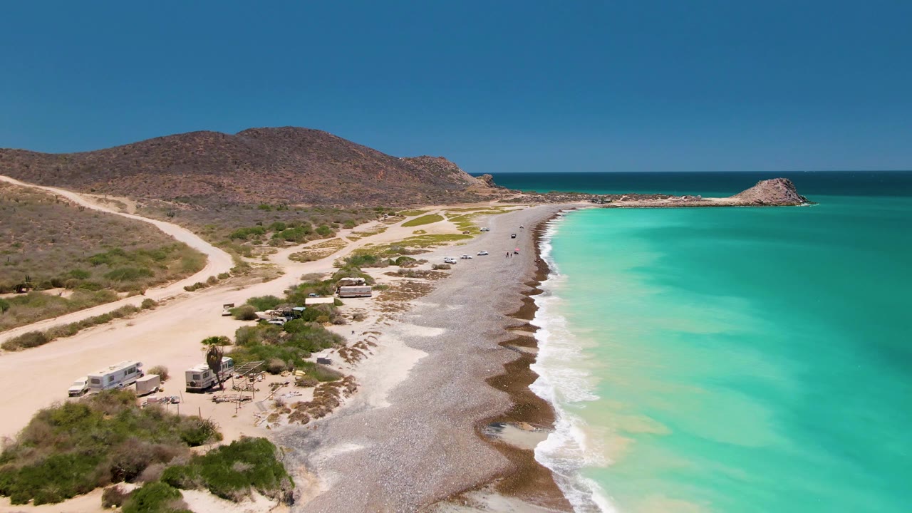 Cabo Pulmo aerial frame with scrubland and vehicles along the sandy road, white surf on the beach, bright turquoise water, brown hills, a small rocky mound offshore, and no visible people.