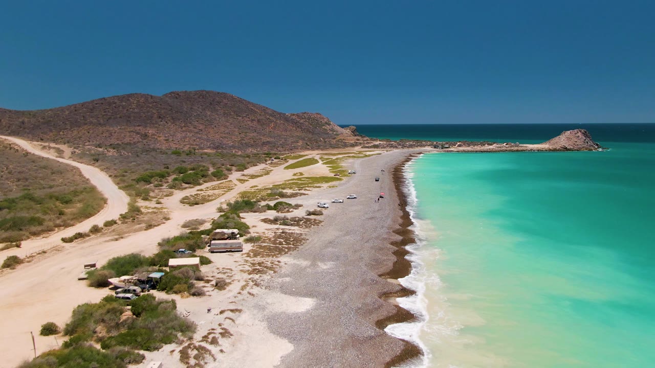 Aerial frame over Cabo Pulmo showing a gray pebbled beach beside turquoise water, dirt tracks at left, small vehicles near beach shelters, distant people, and low brown hills beyond the shore.