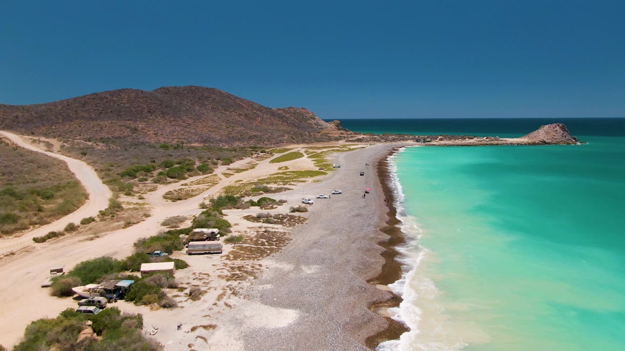 Aerial coastline with gray beach, blue-green water, dirt tracks, cars, and low hills.