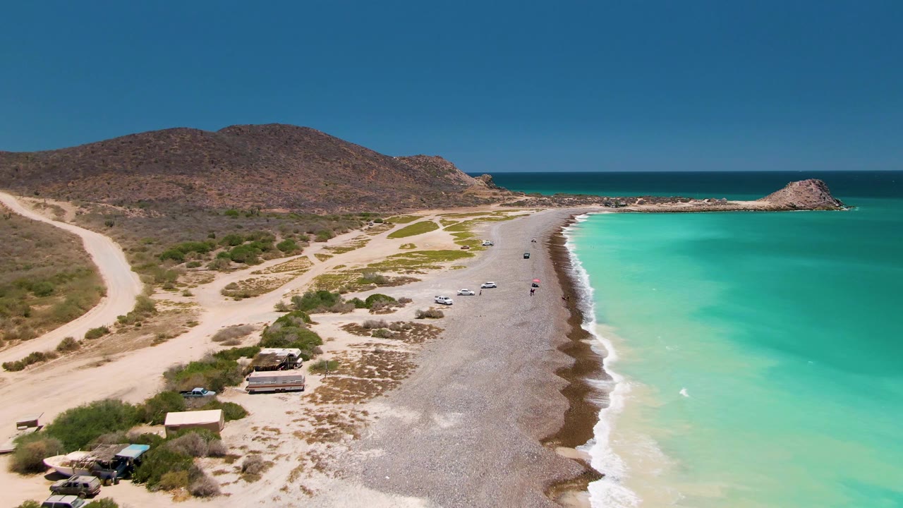 Aerial view over Cabo Pulmo with more of the curved shoreline visible, white foam tracing the water edge, vehicles spaced along the pebbled beach, people near the surf, and a rocky point at the far right.