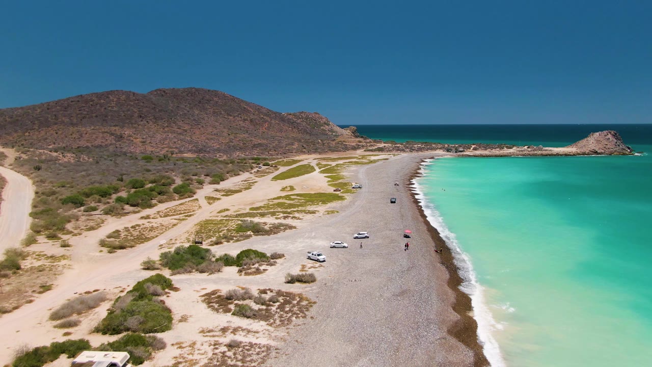Aerial view over Cabo Pulmo, Mexico, showing a sandy beach road beside turquoise water, parked or moving vehicles, low desert shrubs, rocky hills, and tiny people near the shore.