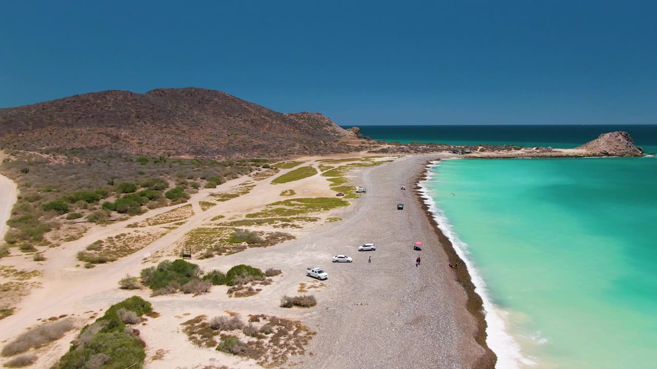 Drone view of vehicles on a beach road with turquoise sea and desert hills.