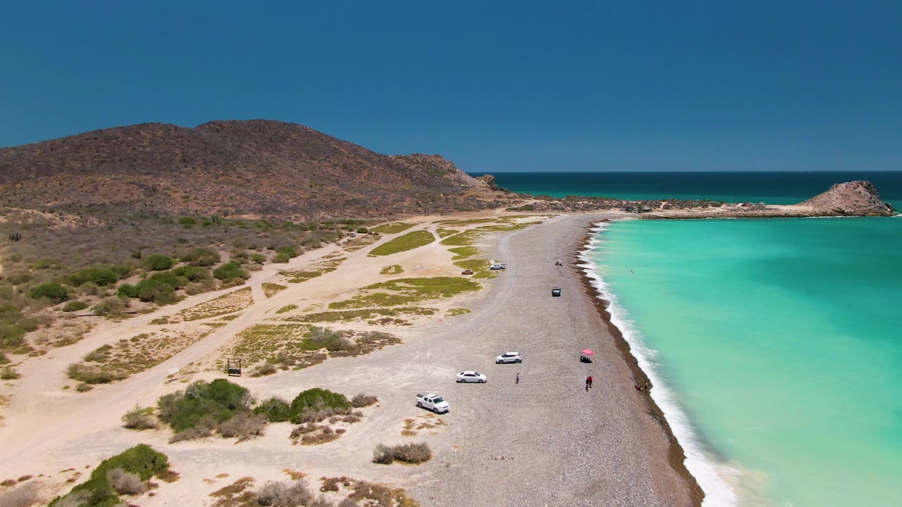 Aerial view in Cabo Pulmo showing a curved pale beach road, scattered cars, a small red umbrella or object near the shore, turquoise water, rocky headland, and dry shrubs.