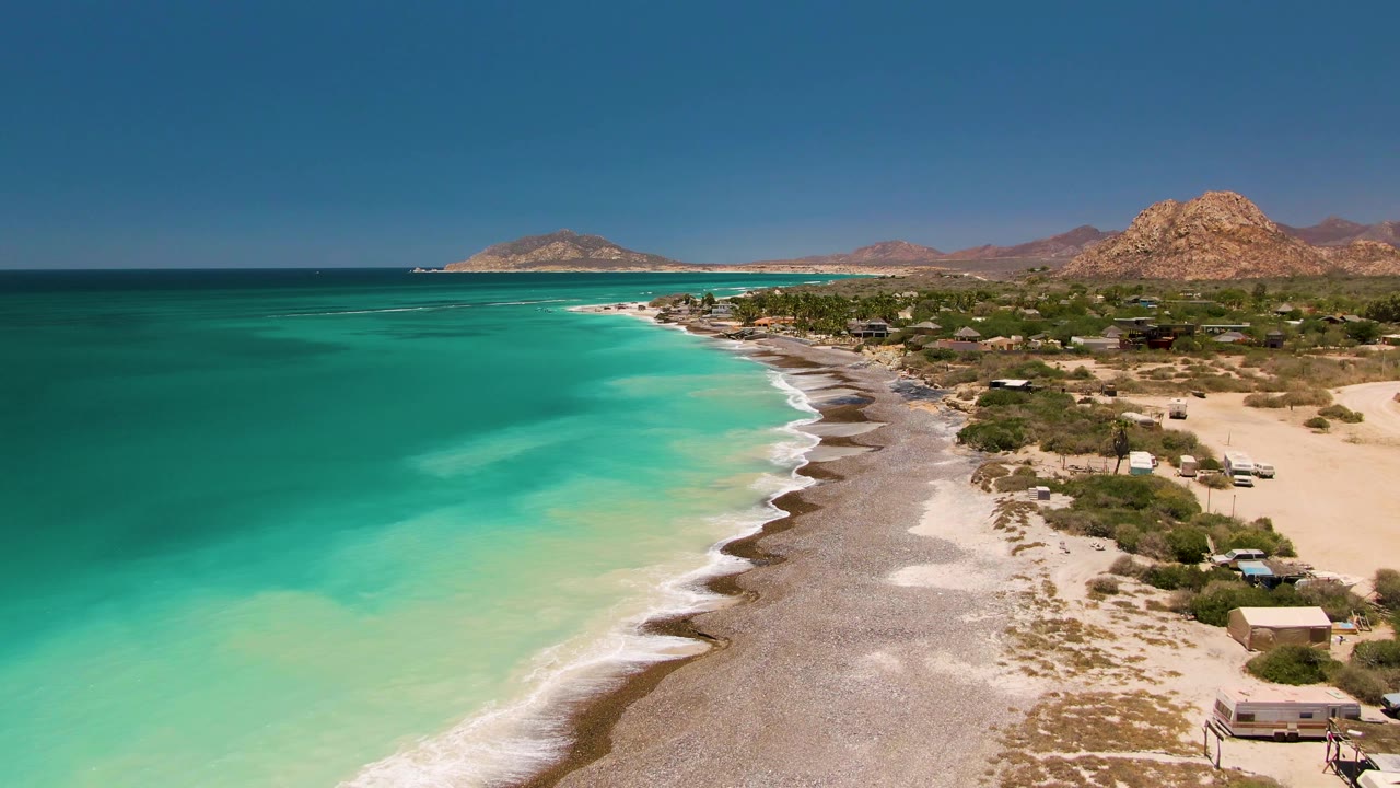 Aerial frame over Cabo Pulmo, Mexico, with turquoise water on the left, a pale surf line, pebbly shore, scattered buildings, dry hills, and no visible people.