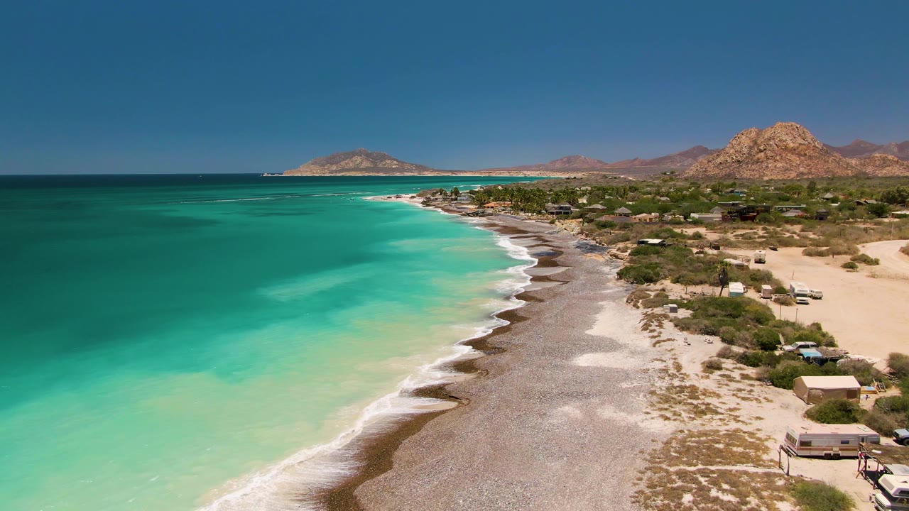 Cabo Pulmo shoreline from above, showing darker offshore water, pale turquoise shallows, a narrow white wave line, sandy lots, low buildings, dry hills, and no visible people.