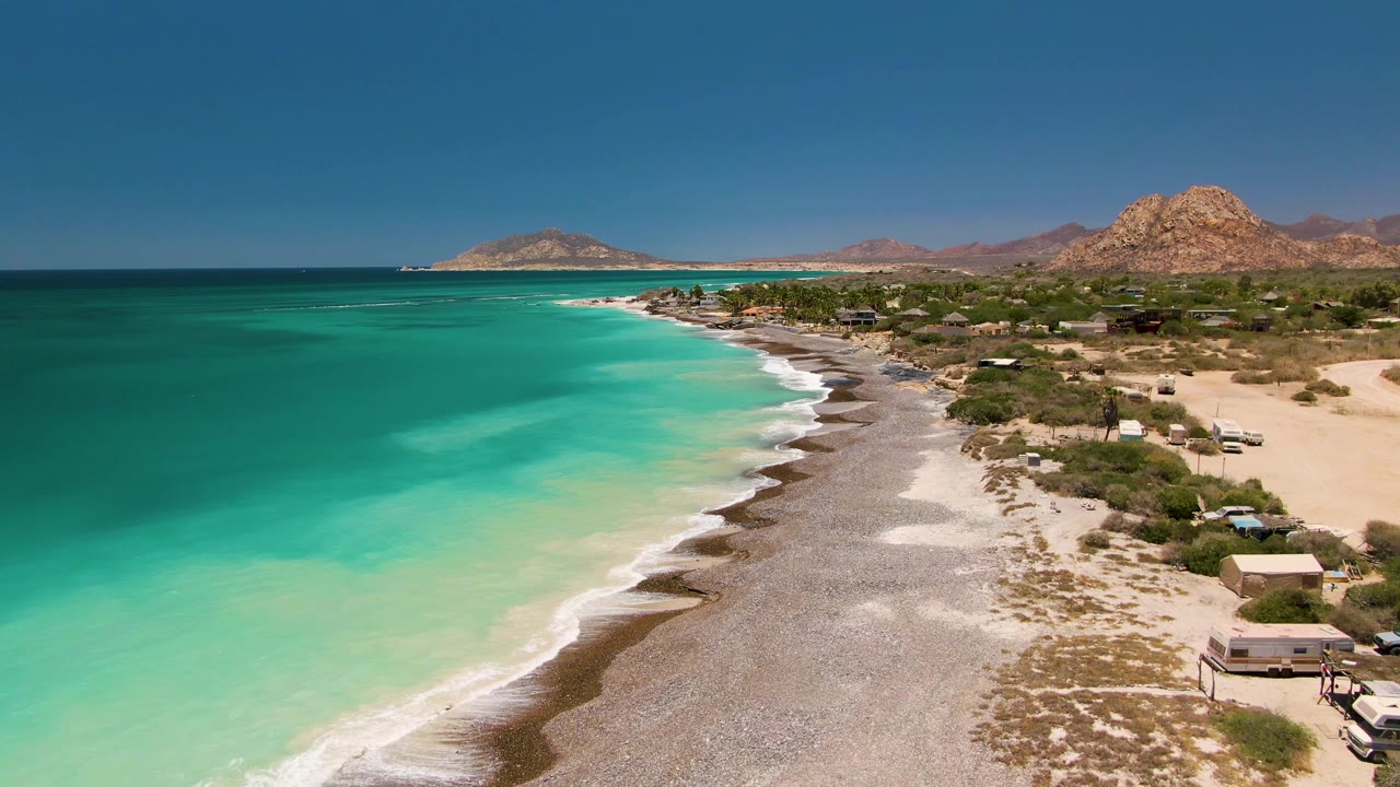 Mid-clip aerial over Cabo Pulmo with turquoise surf washing along the pebbly beach, small structures on the sandy side, rocky hills in the distance, and no visible people.