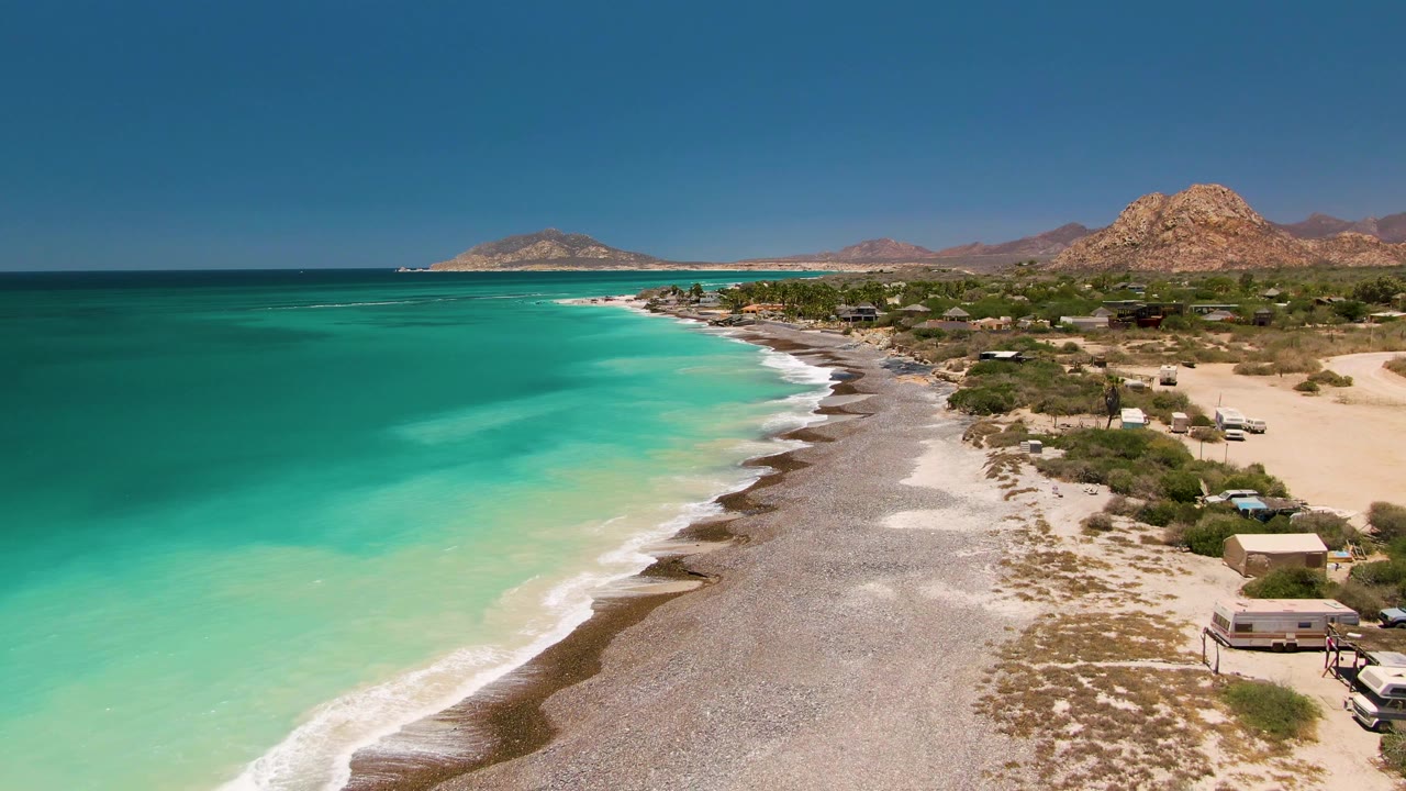 Wide drone view of a pebbly beach with turquoise ocean and desert hills.
