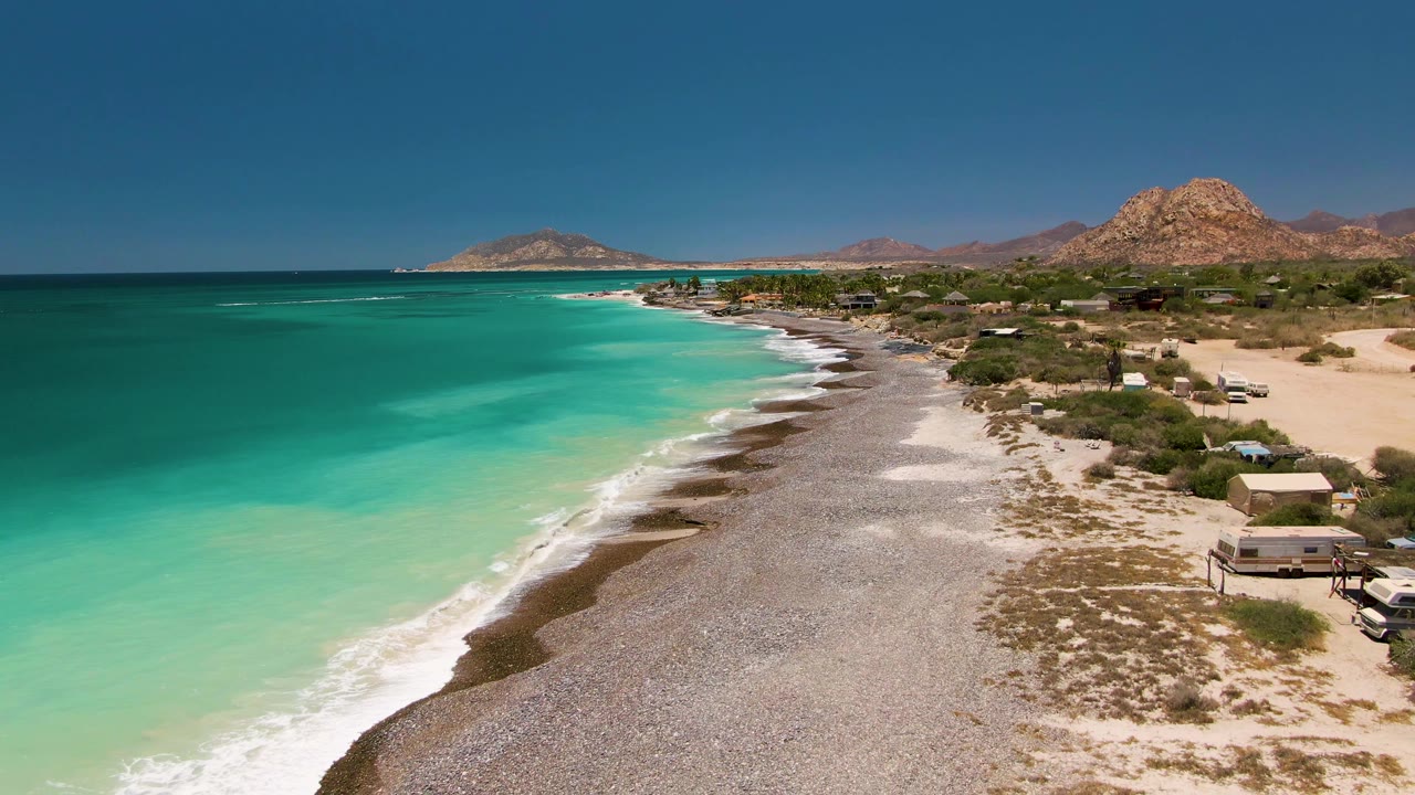 Aerial view of Cabo Pulmo coastline with turquoise water on the left, a gray pebbled shore through the center, sandy scrubland and small structures on the right, and no visible people.